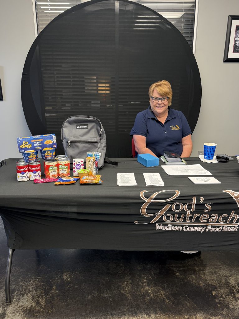 A woman representing God's Outreach is sitting at a table in Richmond Car and Truck Center giving information about the Backpack Program in Madison County. 