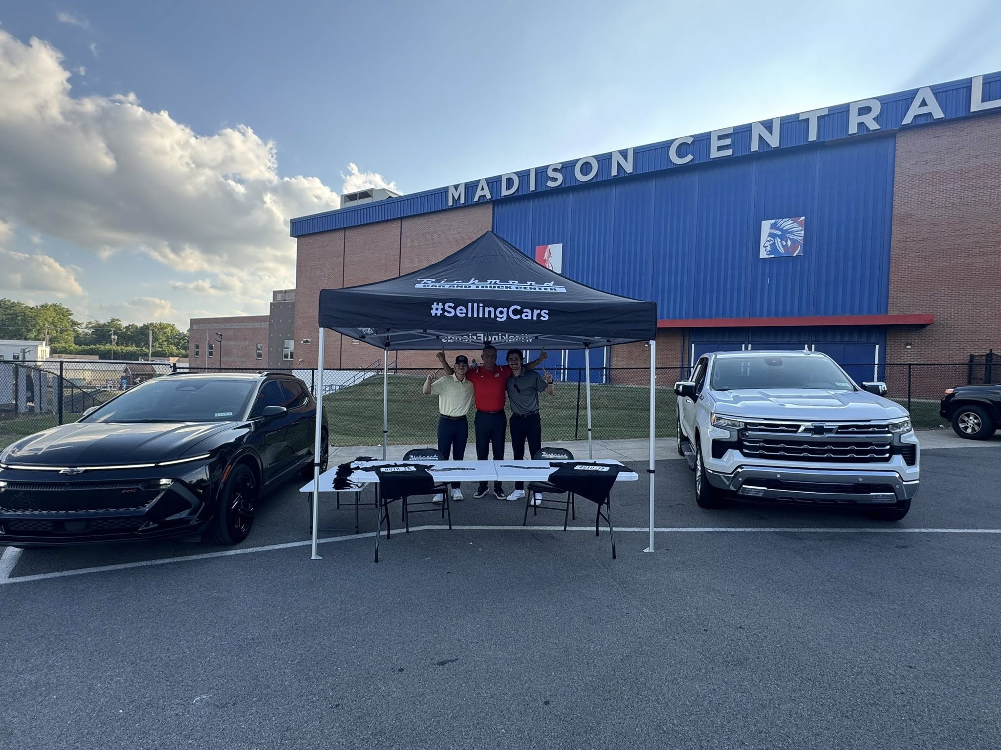 From Left: a Black Chevrolet Blazer EV is parked while Bradyn, Mike, and Jason are underneath Richmond Car and Truck Centers Event Tent in front of madison central high school. A silverado is parked to the right of the tent.